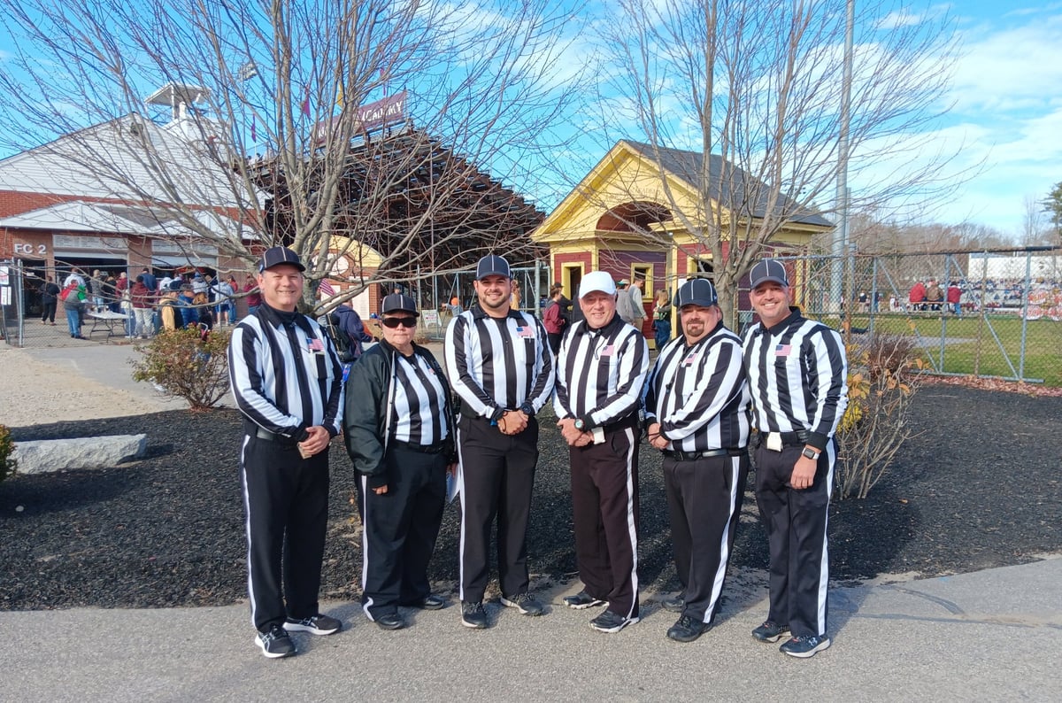 Five sports referees in striped uniforms standing together outdoors in front of a yellow building.